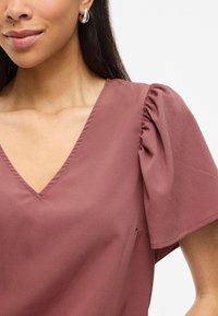 Blusa a V in un tenue mauve con maniche corte a sbuffo, morbida al tatto, caratterizzata da dettagli di cucitura sottili e una vestibilità rilassata.