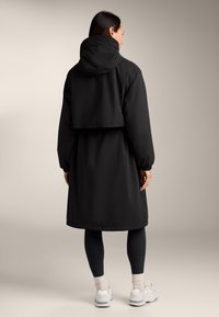 Parka noire à capuche, résistante à l'eau, avec un design court, des poignets élastiques et une texture lisse, portée par-dessus des leggings noirs et des baskets.