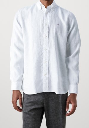 Man draagt een wit overhemd met lange mouwen en knopen, met een klein logo op de borst, en donkergrijze broek.