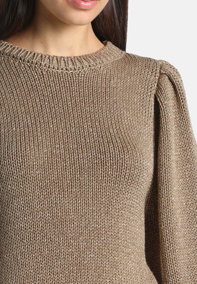 Gros plan d'une femme portant un pull en tricot beige scintillant avec un col rond et un léger détail froncé aux épaules.