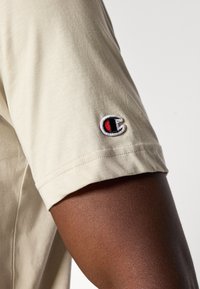 Beige bomulds t-shirt med korte ærmer, der har et lille broderet logo på ærmet i rød, sort og hvid. Glat tekstur.