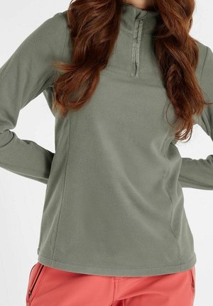 Femme portant un pull polaire vert olive à manches longues avec une fermeture éclair partielle à l'avant et un pantalon corail, les mains posées sur les hanches.