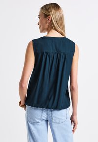 Blouse sans manches de couleur bleu sarcelle foncé avec un détail plissé au dos et un col rond. Tissu lisse et léger ; coupe légèrement ample.