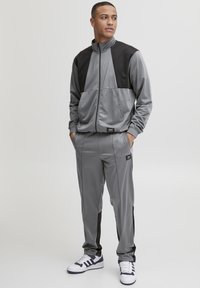 Chándal gris con acentos negros, chaqueta con cuello alto y cremallera, tela ligera, pantalones ajustados, puños elásticos y detalles de la marca.