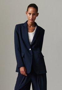 Granatowa marynarka o dopasowanym kroju, z zapięciem na jeden guzik, klapami z wycięciem i przednimi kieszeniami, zestawiona z białym topem na ramiączkach. Gładka faktura.