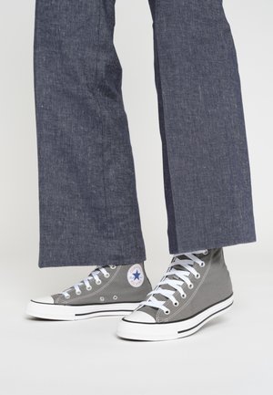 Persona che indossa pantaloni larghi blu scuro e sneakers Converse alte grigie con lacci bianchi, in piedi su una superficie chiara.