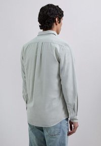 Camisa verde claro de botones con mangas largas, pliegue en la espalda y dobladillo redondeado, hecha de una tela suave. Llevada con jeans azul claro.