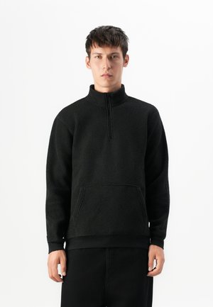 Young man wearing a black fleece pullover with a half-zip and front pocket, standing against a plain white background.