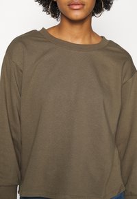 Top de algodón verde oliva de manga larga con un escote amplio, diseño ligeramente corto y textura suave. Sin patrones ni elementos metálicos destacados.