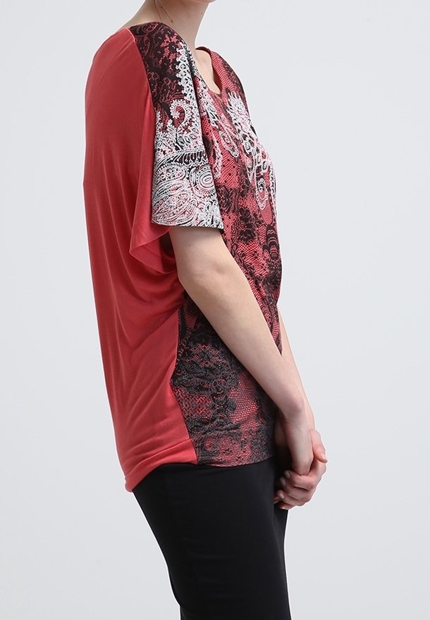 Femme portant un haut à manches courtes rouge et noir avec des motifs cachemire, debout de profil, les mains jointes devant elle.