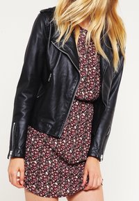 Svart skinnbikerjacka med silverfärgad dragkedja, bärs över en blommönstrad klänning. Har en åtsittande passform och strukturerad yta.