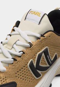 Κοντινό πλάνο χρυσού πλεκτού αθλητικού παπουτσιού με λευκά κορδόνια, εκρού γλώσσα και μεγάλο ασπρόμαυρο λογότυπο "KARL" στο πλάι.