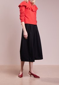 Pull rouge à volants, jupe midi noire et escarpins rouges à bout pointu. La tenue présente une coupe décontractée avec un tissu texturé et un ourlet droit.