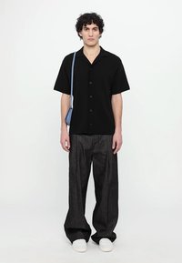 Cămașă neagră cu mâneci scurte și butoni, asociată cu pantaloni largi închiși la culoare și adidași albi. O geantă de umăr bleu deschis adaugă un strop de culoare.