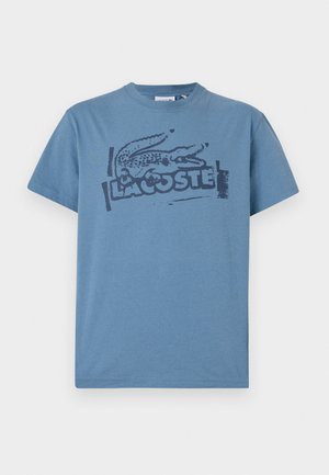 Camiseta de algodón azul con un logo de cocodrilo impreso en negro y el texto "LACOSTE". Cuello redondo, mangas cortas, corte relajado.