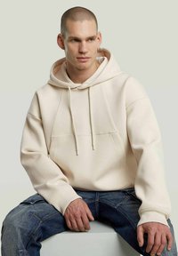 Crème-kleurige hoodie met een trekkoordcapuchon, geribbelde manchetten en een voorkantzak. Gelaagd stof, comfortabele pasvorm, gedragen met donkere spijkerbroek.