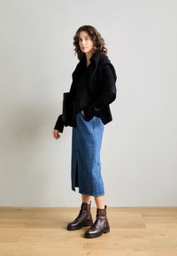 Jeune femme aux cheveux bouclés portant un pull noir sur une chemise noire, une jupe midi en denim bleu et des bottes à lacets marron, debout sur un sol en bois.