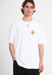 Νεαρός άνδρας με ανοιχτά καφέ μαλλιά, που φοράει ένα λευκό T-shirt με ένα μικρό μαύρο και πορτοκαλί λογότυπο φλόγας στο στήθος και ένα ασημένιο κολιέ.