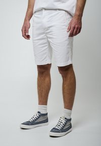 Pantalones cortos de algodón blancos con cierre de botón, bolsillos laterales y dobladillo cosido. Combinados con zapatillas altas grises y calcetines tobilleros blancos.