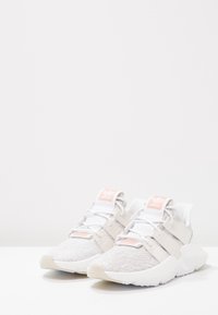 Vaaleanharmaat urheilukengät, joissa on teksturoitu neulososa, valkoisia yksityiskohtia ja vaaleanpunainen brändäys. Liukas design, jossa on elastiset hihnat ja pehmeät pohjat.