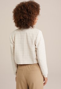 Veste courte de couleur crème avec un tissu texturé et des manches longues. Associée à un pantalon élastique de couleur marron clair avec des poches arrière.