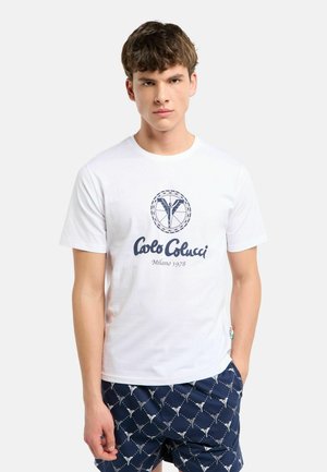 Jonge man met een wit Carlu Colucci T-shirt en donkerblauwe korte broek met een herhalend wit patroon, staand met één hand in zijn zak.