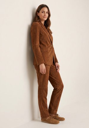 Kvinde med langt brunt hår læner sig op ad beige væg iført en brun ruskind blazer, matchende bukser og brune snørestøvler.