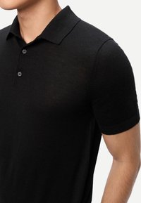 Homme portant un polo en maille noir à manches courtes avec deux boutons, col visible, sur fond blanc.