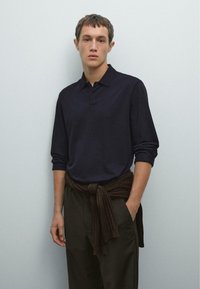 Junger Mann, der ein dunkelblaues Langarm-Polohemd, dunkle Hosen und einen braunen Pullover, der um die Taille gebunden ist, trägt und vor einer hellgrauen Wand steht.