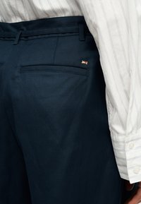 Κοντινό πλάνο τσέπης κοντά navy μπλε ραμμένων παντελονιών με ένα μικρό ορθογώνιο μεταλλικό λογότυπο και μέρος του μανικιού λευκού ριγωτού πουκάμισου.