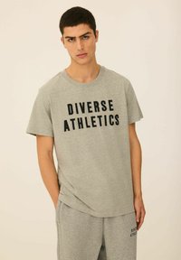 Szara koszulka z napisem "DIVERSE ATHLETICS" w kolorze czarnym, zestawiona z pasującymi szarymi spodniami dresowymi. Wykonana z miękkiego materiału o luźnym kroju.