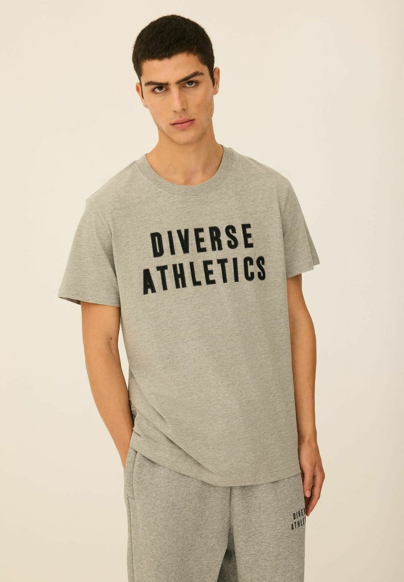 Szara koszulka z napisem "DIVERSE ATHLETICS" w kolorze czarnym, zestawiona z pasującymi szarymi spodniami dresowymi. Wykonana z miękkiego materiału o luźnym kroju.