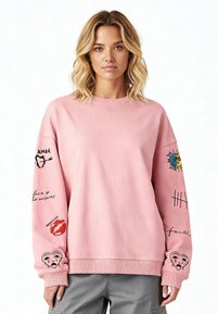 Ružičasti dukserica izrađena od mekog materijala, s raznim šarenim vezovima na rukavima, uključujući motive srca i sunca.