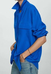 Camisa azul abotonada con cuello doblado, dos bolsillos en el pecho y mangas remangadas, hecha de un tejido liso y ligero.