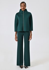 Ensemble vert en deux pièces comprenant un haut zippé ajusté avec un col haut et un pantalon à jambe large. Fabriqué en tissu lisse et texturé.