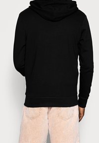 Persona vestida con una sudadera con capucha negra y pantalones de pana color beige claro, mostrada de espaldas contra un fondo liso claro.