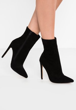 Czarne zamszowe botki na szpic z bocznym zamkiem błyskawicznym i wysokim obcasem typu szpilka, noszone na gołych nogach na jednolitym tle.