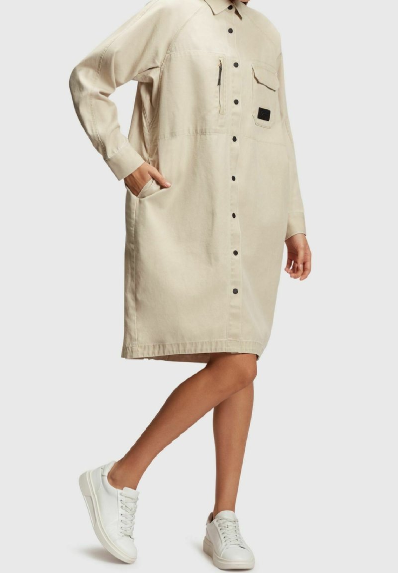 Robe boutonnée beige à manches longues, avec poche plaquée et détail zippé. Portée avec des baskets blanches, elle présente une coupe décontractée et un design minimaliste.