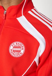 Rød sportsjakke med hvite detaljer, med høy krage, glidelås og FC Bayern Munchen-logo i sirkulært design.