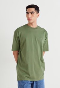 Ung mand med kort krøllet hår iført en overdimensioneret olivengrøn T-shirt og blå jeans, som kigger til venstre.