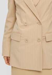 Béžové sako s jemným proužkem, dvouřadé s čtyřmi knoflíky a kapsami s klopami, nošené přes sladěnou béžovou sukni.