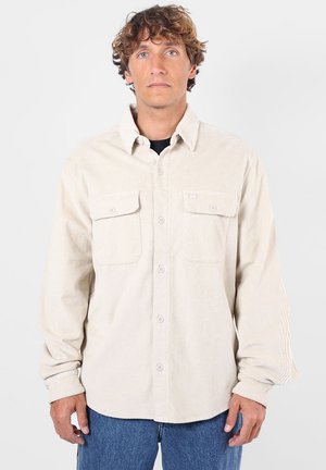 Homme aux cheveux bouclés portant une chemise en velours côtelé beige à boutons par-dessus un sous-vêtement noir et un jean bleu, debout devant un fond uni.