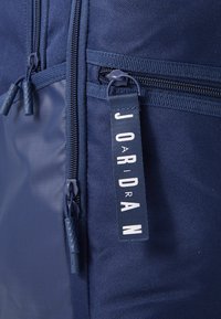Sötétkék hátizsák texturált anyaggal, több cipzárral és "JORDAN" felirattal ellátott címkével. Tartós hardverrel és sima részletekkel rendelkezik.