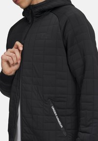 Chaqueta negra acolchada con capucha, que presenta un cierre de cremallera, puños elásticos y un logo de Under Armour en el frente.