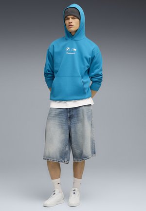 Turkis hættetrøje med "Motorsport" logo, parret med lyseblå denim shorts og hvide sneakers. Hvide crew-sokker.