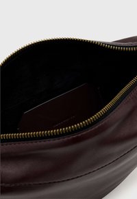 Bordeauxrode leren tas met een gouden ritssluiting en zwarte binnenkant. Heeft een gladde textuur en een klein donker kaartje zichtbaar aan de binnenkant.