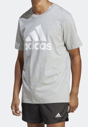 Personne portant un t-shirt gris Adidas avec un grand logo et un short Adidas noir, debout devant un fond uni.