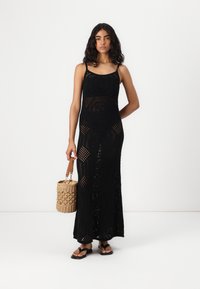 Μαύρη κροσέ μέγιστη φόρμα με λουριά τύπου spaghetti, που διαθέτει γεωμετρικά σχέδια. Συνδυασμένη με μια πλεκτή τσάντα και επίπεδες σανδάλια.