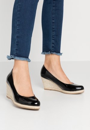 Füße, die dunkelblaue Jeans mit ausgefranstem Saum und schwarze Lack-Wedges-Espadrilles mit geflochtenen Hanfsohlen tragen.