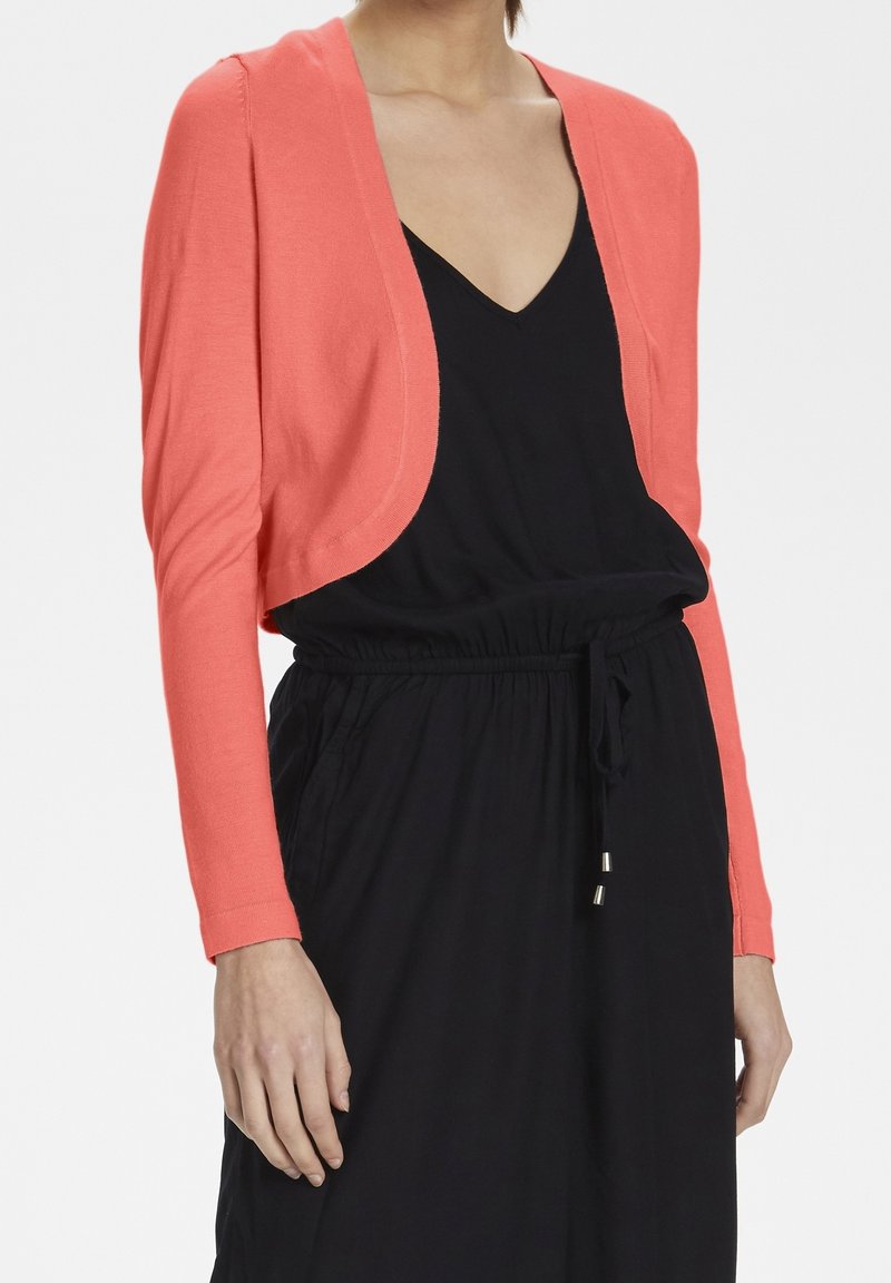 Femme portant un cardigan court corail sur une robe noire sans manches à col en V avec taille à cordon, debout devant un fond uni.